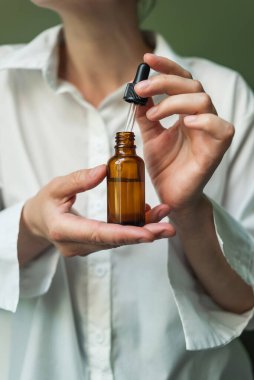 Yeşil arka plandaki bir kadının elinde pipet olan kahverengi bir şişedeki temel yağ. Bir şişe yağ ve pipetle kadın eli. Güzellik küresi.