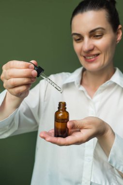 Beyaz gömlekli, gülümseyen bir kadın, elinde bir şişe yağ tutuyor ve bir pipetten damlatıyor. Beyaz bir kadının ellerinde pipeti olan bir esans.