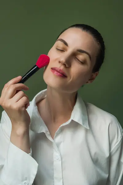 stock image Middle-aged European women holds a pink makeup brush to her face. Face makeup. A woman with a makeup brush on a green background