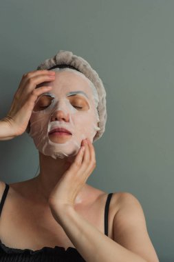 Kumaş maskeli kadın cilt bakımı yapıyor. Gri bir arka planda yakın plan portresi. Güzellik endüstrisi, kişisel bakım..