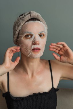 Kumaş maskeli kadın cilt bakımı yapıyor. Gri bir arka planda yakın plan portresi. Güzellik endüstrisi, kişisel bakım..