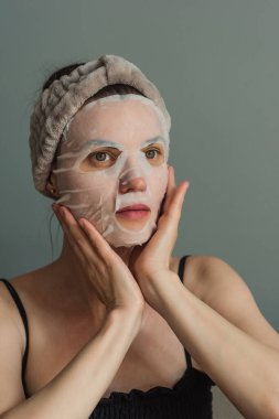 Kumaş maskeli kadın cilt bakımı yapıyor. Gri bir arka planda yakın plan portresi. Güzellik endüstrisi, kişisel bakım..