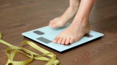 Beyaz zemin terazileri ve üzerinde duran kadın ayakları. Vücut değişimlerini ölçmek için yakınlarda duran bir terzi ya da mezura şeridi. Sağlıklı yaşam tarzı ve vücut ağırlığı takibi kavramı