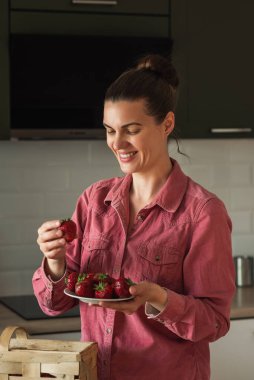 Gülümseyen olgun bir kadın, parlak bir mutfakta, masasında bir sepetin yanında çilek tutar. Yazın reçel, salata yapar ve taze böğürtlen yerim. Sağlıklı eko-çiftlik, organik ev yapımı yiyecekler.