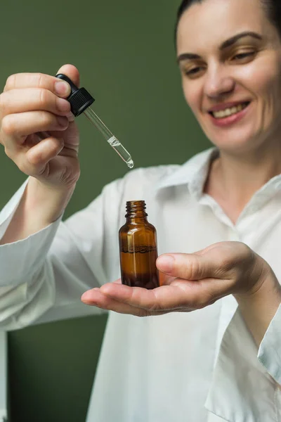 Gülümseyen bir kadın kahverengi bir yağ şişesini elinde ve pipetle tutuyor. Güzellik küresi için esans - yüz için, saç için, deri için