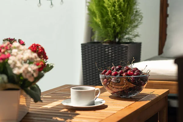 Büyük bir vişne kasesi ahşap bir masanın üzerinde, bir fincanın yanında duruyor. Güneşli bir yaz gününde çiçekler ve meyvelerle dolu rahat bir balkon..