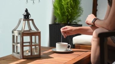 A woman is sitting on a terrace with wooden furniture and brewing tea from a tea bag in boiling water. A woman drinks tea on the balcony