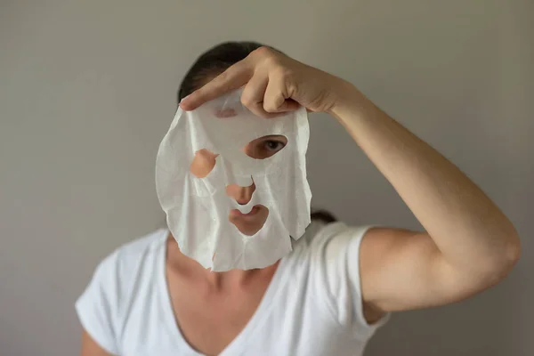 A woman with a fabric cosmetic mask in her hands, preparing for skin care. Close-up portrait on a gray background. The beauty industry, self-care.