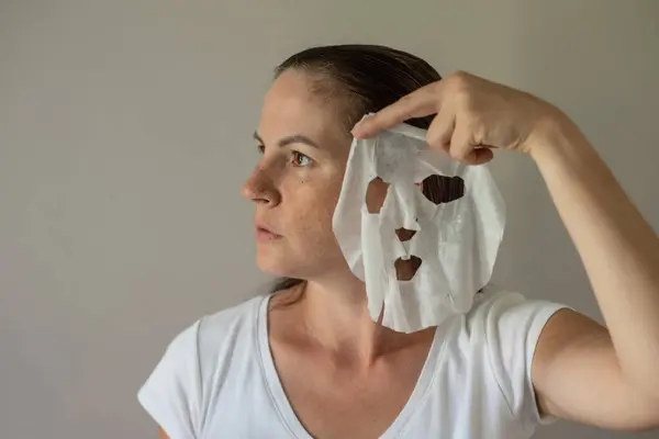 A woman with a fabric cosmetic mask in her hands, preparing for skin care. Close-up portrait on a gray background. The beauty industry, self-care.