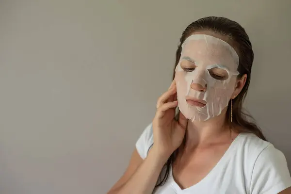 woman with a fabric cosmetic mask on her face, does skin care. Close-up portrait on a gray background. The beauty industry, self-care.