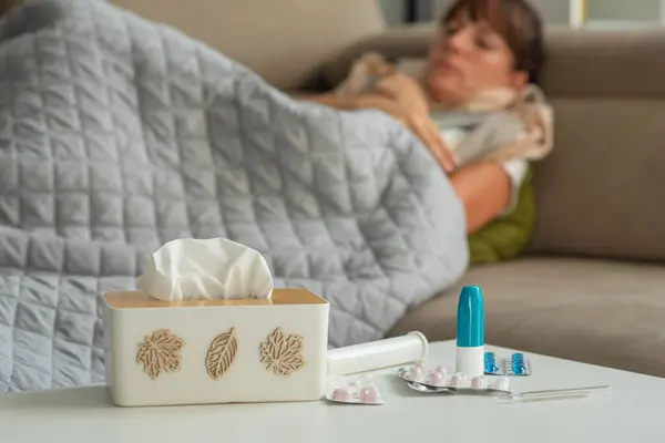 Cold medicines and paper napkins are on the table, a woman with signs of flu and colds is lying on the background. Focus on medications