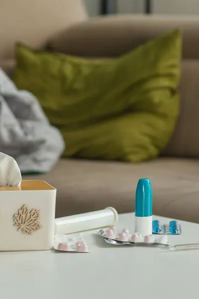 Cold medicine on the background of a sofa with pillows and a blanket. Tablets, spray and thermometer are on the table against the background of a place to relax