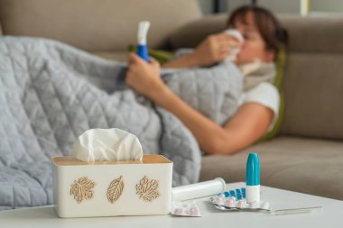 Soğuk algınlığına karşı ilaçlar ve kağıt peçeteler masada, grip ve soğuk algınlığı belirtileri gösteren bir kadın arka planda uzanıyor ve solunum yapıyor. İlaçlara odaklan.