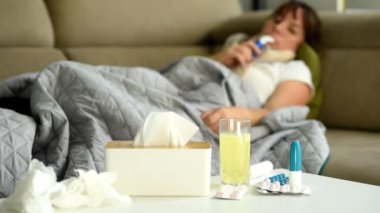 İlaçlar ve tıbbi malzemeler masanın üzerine serildi. Bir kadın kanepede uzanıyor ve solunum yapıyor. Hastalık ve evde sağlık sorunu