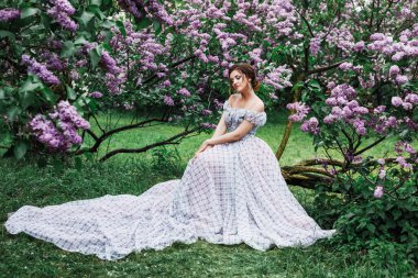 Kıvırcık saçlı, uzun açık renk elbiseli ve taç giymiş güzel bir esmer kadının bahar portresi, leylak çiçeklerinin dallarında oturuyor.
