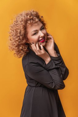 Sarı arka planda siyah tişörtlü, kıvırcık saçlı, güzel, yaşlı bir kadının şık stüdyo resimleri.