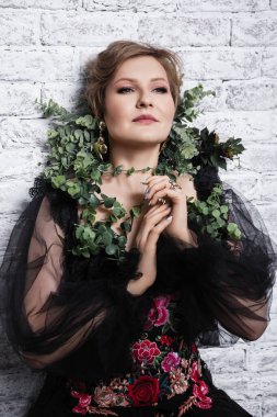 Tüylü, korseli ve yeşil dallı siyah bir gece elbisesi içinde güzel bir kadın tuğla bir duvara yaslanmış oturuyor.