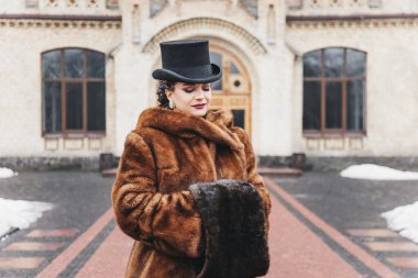 Eski bina, kürk manto, saç örgüsü, akşam makyajı, mavi gözler, kar, eski bina yakınlarında esmer ve silindir şapkalı güzel bir kadının eski moda portresi.