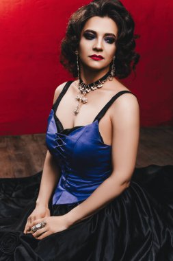 Gece makyajlı, kıvırcık saçlı, siyah ve mor korse elbiseli, kırmızı dudaklı, güzel esmer kadın, ahşap zeminde kırmızı bir duvarın yanında oturuyor.