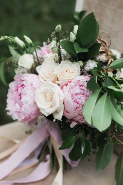 Krema güllü ve pembe şakayıklı bir düğün buketi çimenlerdeki bir sandalyede duruyor, yeşil yapraklı ve dutlu dallar, saten kurdeleler