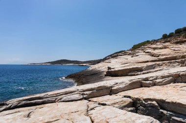 Adanın deniz manzarası, kayalık plaj, yeşil tepeler ve dağlar, mavi deniz ve açık gökyüzü.