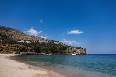 Yaz tatilinde adada deniz manzarası, altın plaj, yeşil tepeler, dağlar ve kayalar, bir yat, mavi ve turkuaz deniz ve berrak gökyüzü.