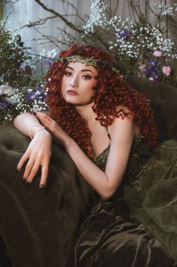 Kanepede oturan çelenkli fantezi kadın. Bahar çiçekleriyle süslenmiş bir oda. Çiçek açmış şehvetli bir dişi. Kıvırcık kızıl saçlı seksi kadın