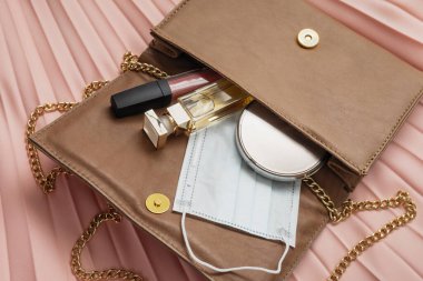 İçinde tıbbi maske, ruj, toz ve parfüm bulunan açık kadın çantası pembe arka planda yatıyor, Coronavirus kadın modası, covid-2019 koruma