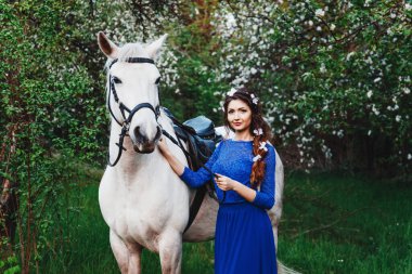 Saçları kıvırcık, akşam makyajlı, mavi elbiseli, saçlarında beyaz çiçekli, ağaçlı, bahçeli, beyaz atlı güzel esmer bir kadının açık hava portresi.