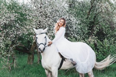 Beyaz uzun elbiseli güzel bir sarışının açık hava portresi ve saçlarında çiçekler beyaz bir at, çiçekler, örgü, saç stili, makyaj...