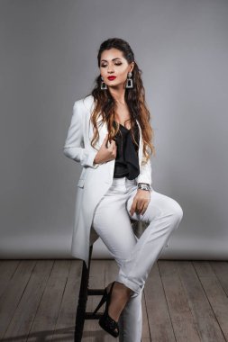 Beyaz takım elbiseli, beyaz tüylü küpeli, kırmızı rujlu, uzun bacaklı, yüksek topuklu, makyajlı, ceketli güzel esmer bir kadının stüdyo portresi.