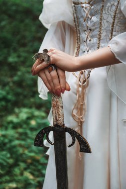 Beyaz ortaçağ kostümü giymiş, yeşil bir ormanda duran bir kartal yüzüğüyle kollarında bir kılıç tutan fantezi prensesi olan bir kadının yakın çekimi.