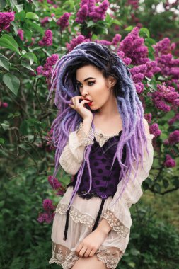 Renkli rastalı seksi genç kadın model, leylağın altında duruyor, bahar moda portresi.