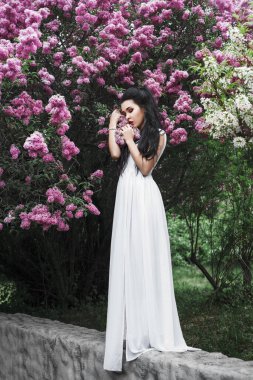 İlkbaharda açık havada güzel, esmer bir kadının portresi leylak çiçekleri arasında, uzun beyaz elbise, siyah saç, akşam makyajı, mücevher, bahçe, çiçek.