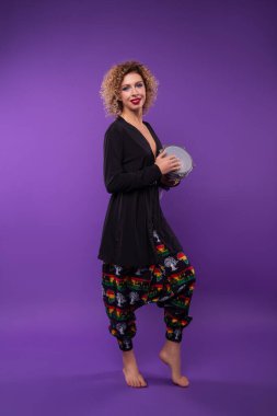 Kırmızı dudaklı, kıvırcık saçlı, siyah gömlekli, Afrika desenli pantolonlu güzel sarışın bir kadın mor bir stüdyo arkasında duruyor ve davul çalıyor.