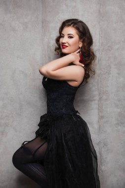Kıvırcık saçlı, gece makyajlı, kırmızı dudaklı, siyah guipure elbiseli ve çoraplı güzel esmer kadın beton bir duvarın yanında duruyor ve gülümsüyor.
