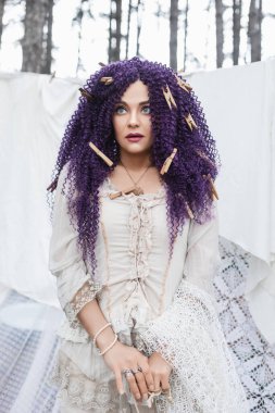 Boho tarzı giyinmiş güzel bir kadın mor kıvırcık saçlı, elbiseleri yıkıyor ve elbiselerini ormanda kurutmak için asıyor, ev hanımı, ev işi, çamaşırhane, doğada yaşam, ekolojik yaşam.