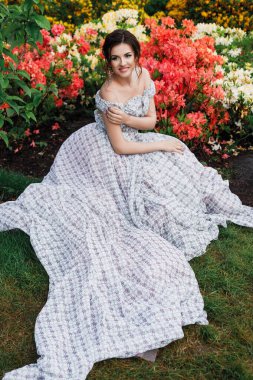 Saçları ve gece makyajı ile güzel bir esmer kadının açık hava portresi, uzun açık renk bir elbise içinde açelya çiçeklerinin içinde duruyor.