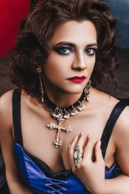 Güzel esmer kadın mor gece makyajı ve kıvırcık saç stili, kırmızı dudaklar, siyah ve mor korse elbise ve büyük elmas haç, yakın çekim