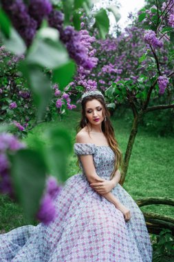 Kıvırcık saçlı, uzun açık renk elbiseli ve taç giymiş güzel bir esmer kadının bahar portresi leylak çiçeklerinin dalları arasında duruyor.