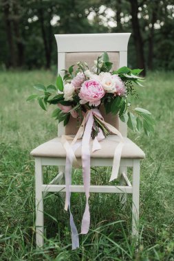 Beyaz peignoir 'li gelin bir düğün buketine krema gülleri ve pembe şakayıklar ile dokunur, yeşil yapraklı dallar ve böğürtlenler, sandalye, çimen, açık hava