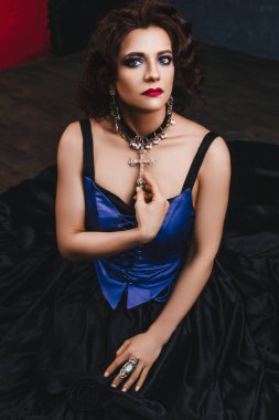 Güzel esmer kadın mor gece makyajı ve kıvırcık saç stili, kırmızı dudaklar, siyah ve mor korse elbise ve büyük elmas haç, yakın çekim