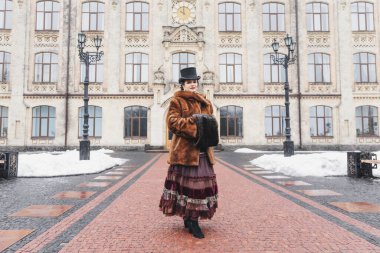 Eski bina, kürk manto, saç örgüsü, akşam makyajı, mavi gözler, kar, eski bina yakınlarında esmer ve silindir şapkalı güzel bir kadının eski moda portresi.