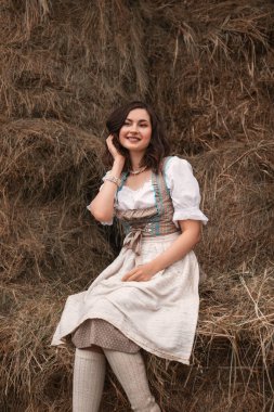 Geleneksel Bavyera kostümü giymiş güzel, genç esmer kadın samanlıkta oturuyor ve gülümsüyor, oktoberfest