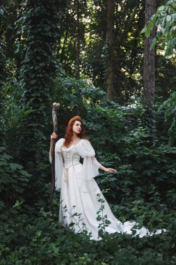 Beyaz bir ortaçağ kostümü içinde, yeşil bir ormanda asası olan güzel kızıl saçlı kadın, fantezi prensesi.