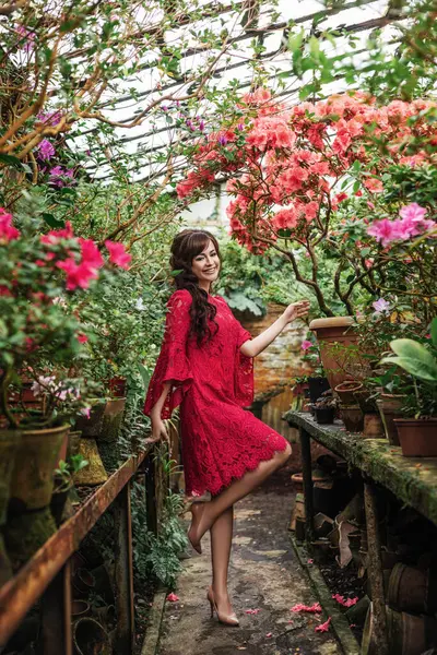 sexy brunette woman in a pink dress is standing in blooming azalea glasshouse, beautiful spring flowers