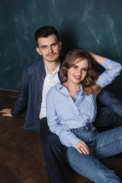 young man and woman in jeans clothes sit together on a wooden floor near a blue wall and smile