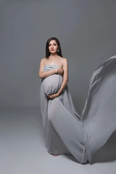 beautiful pregnant woman in flying transparent fabric stands in studio