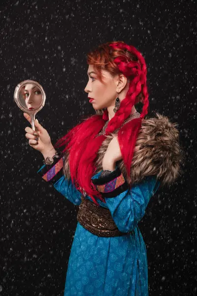 Kırmızı saçlı, kürk pelerinli, mavi elbiseli, elinde aynayla kar yağarken sırtını kameraya dayayıp yansımasına bakan güzel bir kadın.