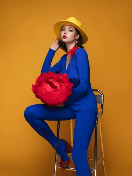 Mavi kostümlü, sarı şapkalı, kırmızı tüylü ve sarı arka planda dev kağıt güllü güzel bir kadının moda stüdyosu portresi.
